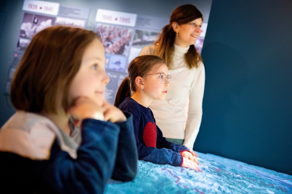 Deux enfants et une adulte observent attentivement une exposition interactive. Les visages des enfants reflètent la curiosité et l'émerveillement, tandis que la femme semble leur expliquer quelque chose. L'arrière-plan montre des panneaux d'information historique, renforçant l'aspect éducatif et captivant de l'expérience.