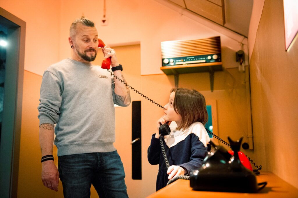 Un homme et une jeune fille interagissent avec des téléphones rétro dans un environnement muséal. L'homme tient un téléphone rouge, tandis que la jeune fille utilise un téléphone noir, simulant une conversation amusante. L'ambiance rappelle une époque ancienne, créant une expérience immersive et éducative.