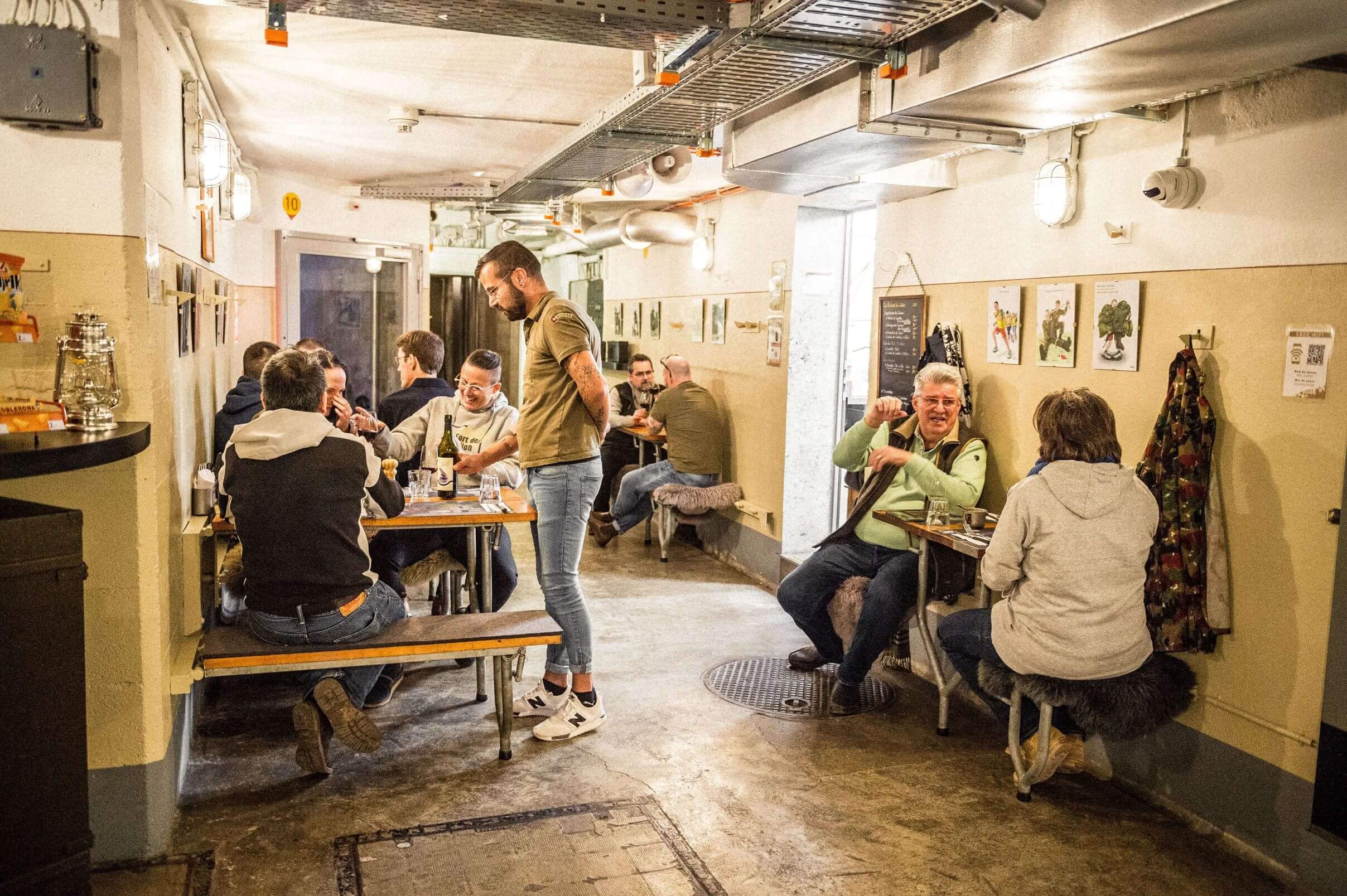 Un groupe de personnes explore un espace muséal interactif, rassemblé autour d'une table couverte d'un drap vert. Ils observent attentivement un objet sur la table, échangeant des commentaires et des sourires. L'environnement est carrelé et éclairé de manière douce, créant une ambiance immersive et éducative.