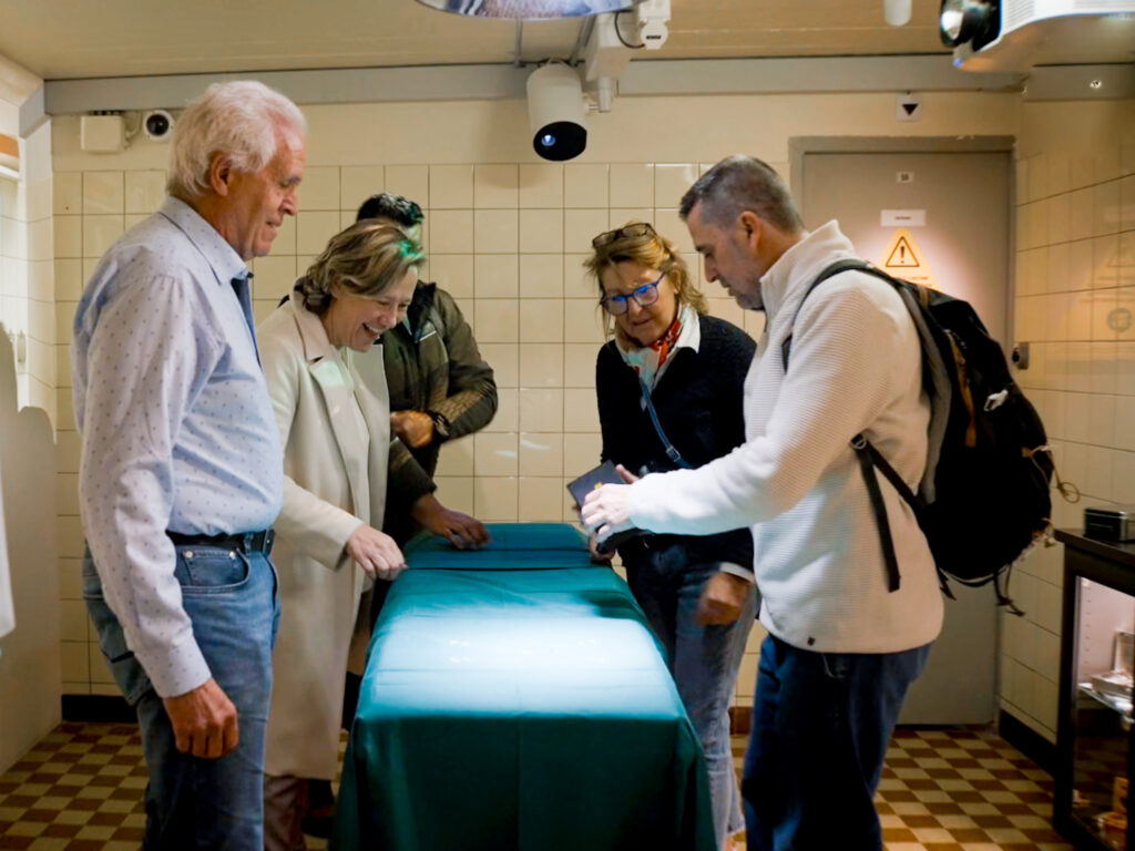 Un groupe de personnes explore un espace muséal interactif, rassemblé autour d'une table couverte d'un drap vert. Ils observent attentivement un objet sur la table, échangeant des commentaires et des sourires. L'environnement est carrelé et éclairé de manière douce, créant une ambiance immersive et éducative.