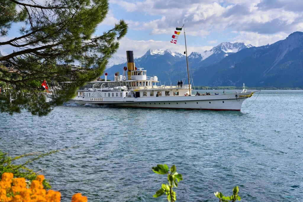 Un bateau à vapeur historique nommé 'La Suisse' navigue paisiblement sur un lac entouré de montagnes majestueuses sous un ciel partiellement nuageux. Le drapeau suisse flotte à l'arrière du bateau, tandis que des passagers profitent de la vue depuis le pont. Des arbres et des fleurs en premier plan ajoutent une touche de couleur naturelle à la scène