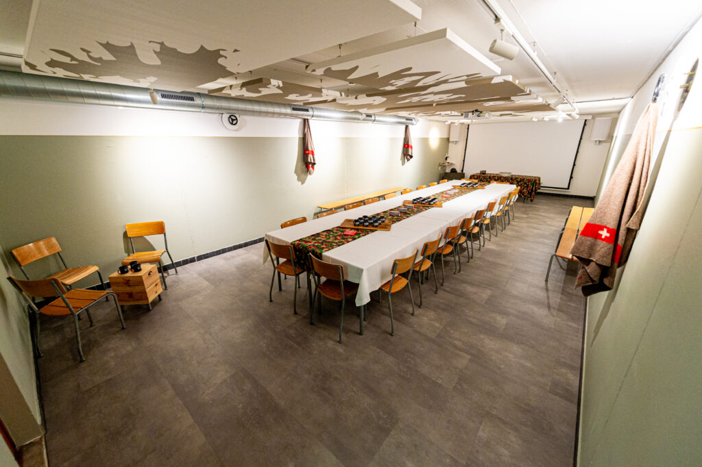 Une grande salle de réunion ou de banquet, aménagée avec une longue table rectangulaire recouverte d'une nappe blanche et décorée de chemins de table aux motifs camouflage. Des chaises en bois sont disposées autour de la table, et des couvertures avec le drapeau suisse sont accrochées aux murs, ajoutant une touche thématique. L'espace est sobre et bien éclairé, prêt pour un événement ou une réunion.