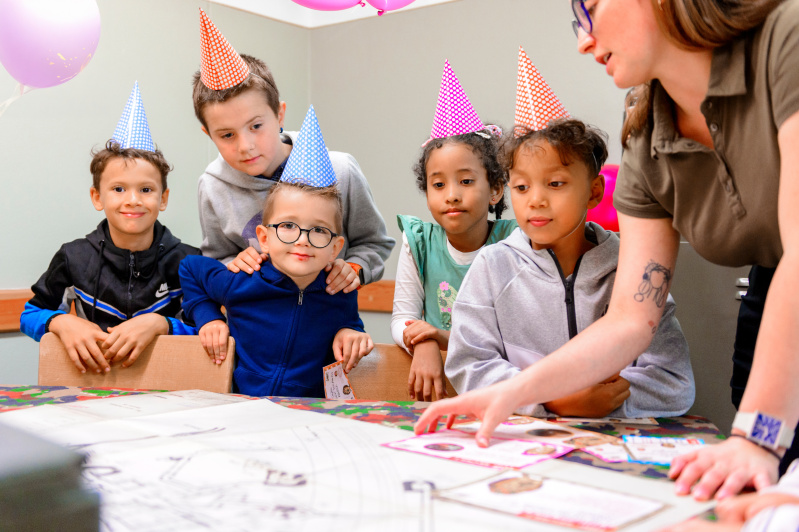 Un groupe d'enfants portant des chapeaux de fête colorés écoute attentivement une animatrice qui leur explique des activités. Ils sont autour d'une table avec des cartes et des feuilles de papier, dans une ambiance d'anniversaire festive avec des ballons roses en arrière-plan. Les enfants semblent curieux et engagés, prêts à participer aux activités.