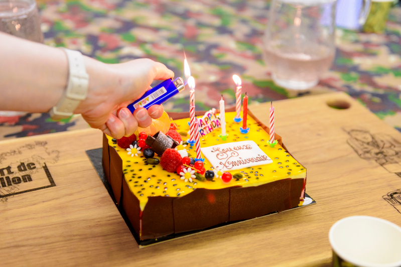 "Un gâteau d'anniversaire coloré avec un glaçage jaune et des décorations de fruits et de fleurs en sucre est en train d'être préparé pour la célébration. Une main allume les bougies avec un briquet, ajoutant la touche finale avant de célébrer. Le message sur le gâteau indique 'Joyeux Anniversaire', et le décor de la table est à motifs camouflage.